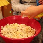 A child spraying margarine on popcorn in a red bowl
