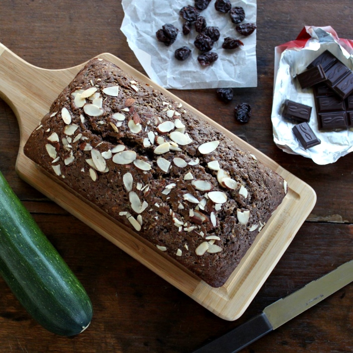 loaf of Zucchini Bread with Dark Chocolate and Dried Cherries on a cutting board with ingredients scattered around