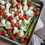 Angled photo of Lemon Pesto Sheet Pan Chicken with Asparagus and Tomatoes sitting on a cutting board with a pot holder