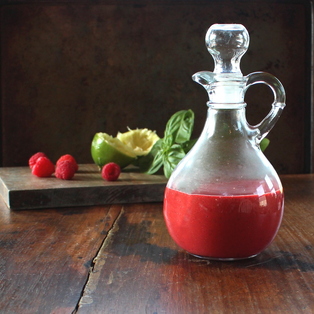 Raspberry Basil Vinaigrette in a cruet in front of cutting board containing the recipe ingredients