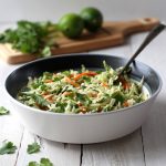 Coleslaw in a white bowl with cilantro and limes in the background