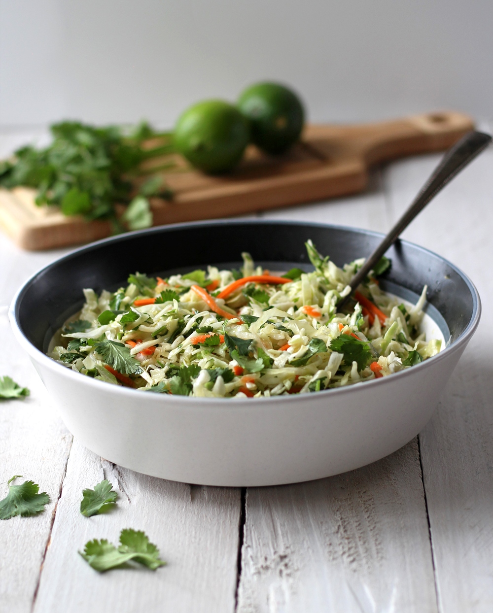Coleslaw in a white bowl with cilantro and limes in the background