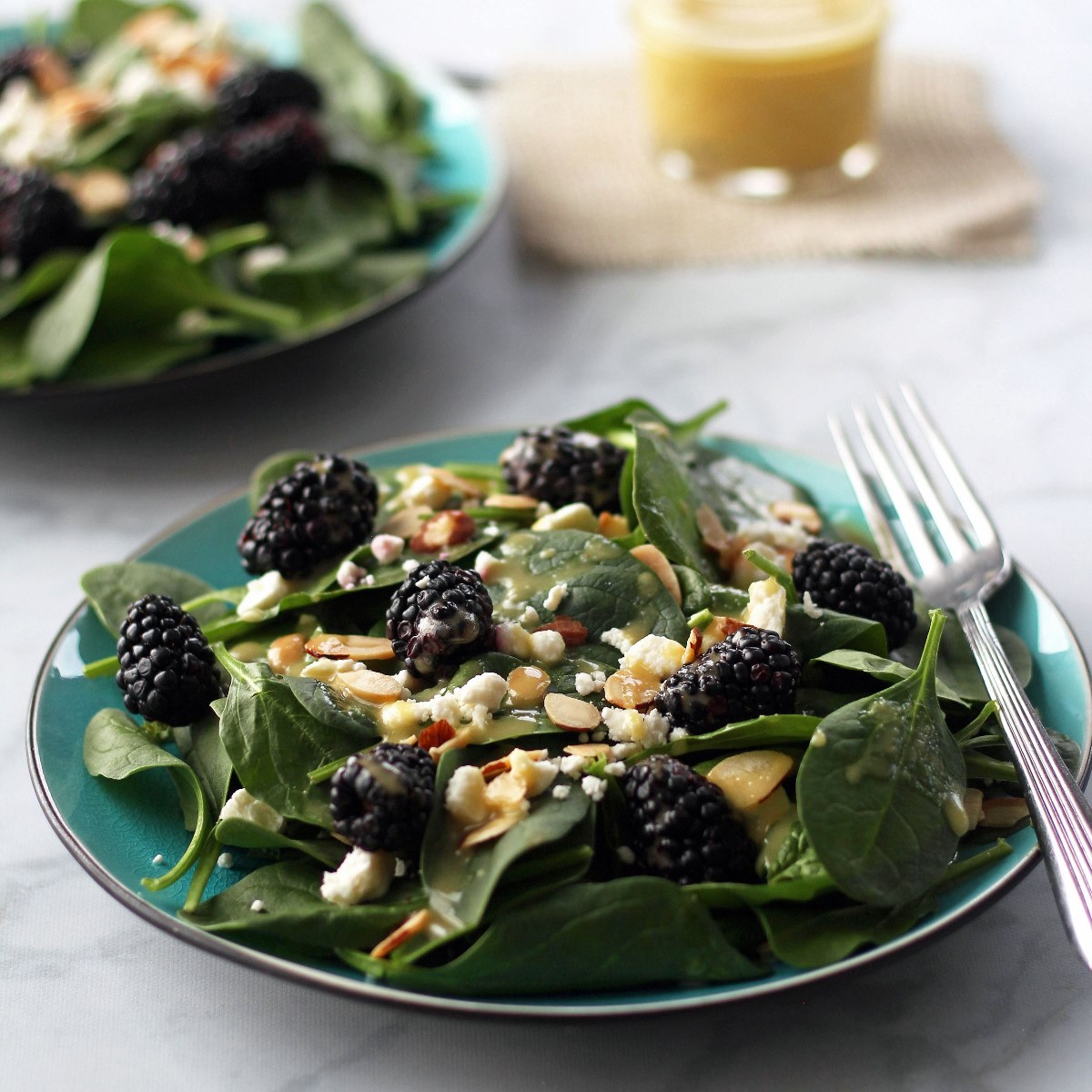 Spinach salad with blackberries, feta cheese, and sliced almonds with dressing on a teal plate and fork. Looking at it from an angle.